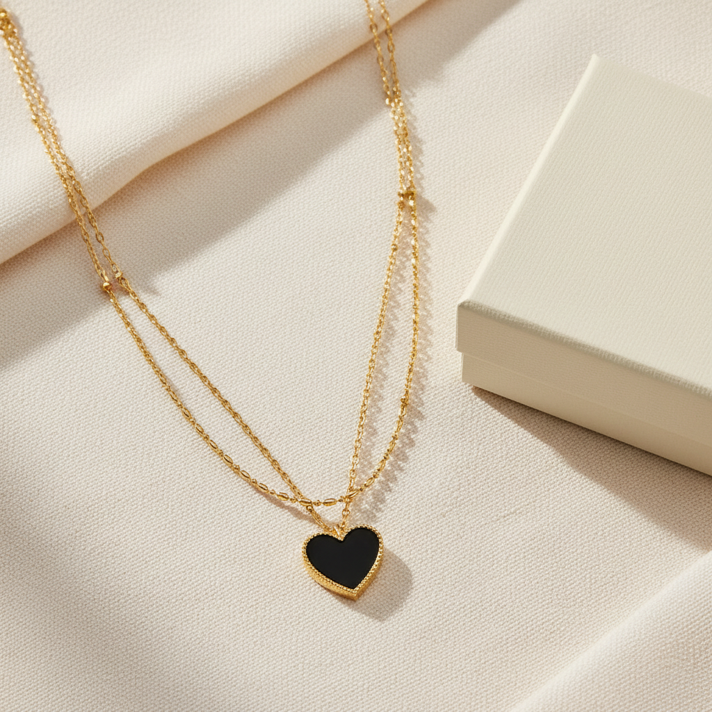 Flat lay of gold layered necklace with onyx black heart pendant and gift box on cream surface